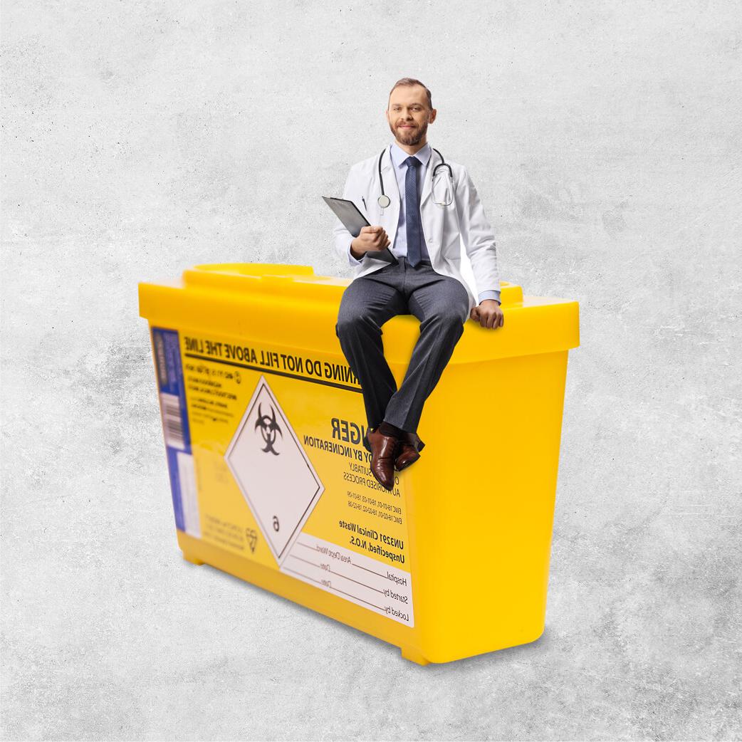 Man wearing a white lab coat and stethoscope sitting on top of an oversized yellow 'sharps' needle bin