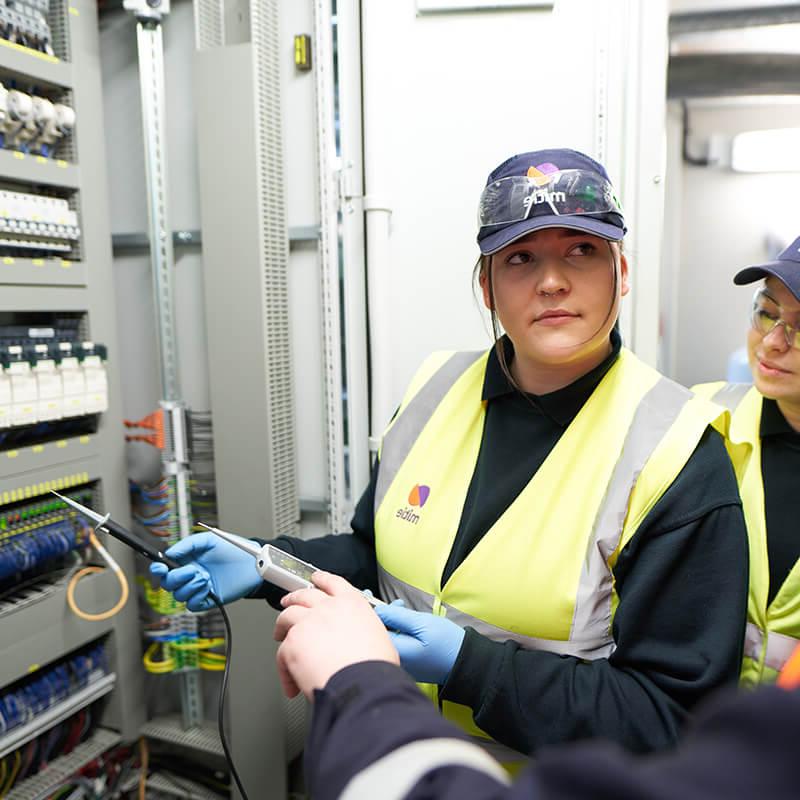 两个女工程师学徒, 穿着Mitie高能见度背心和帽子, in front of a wall of electrical wires and fuses